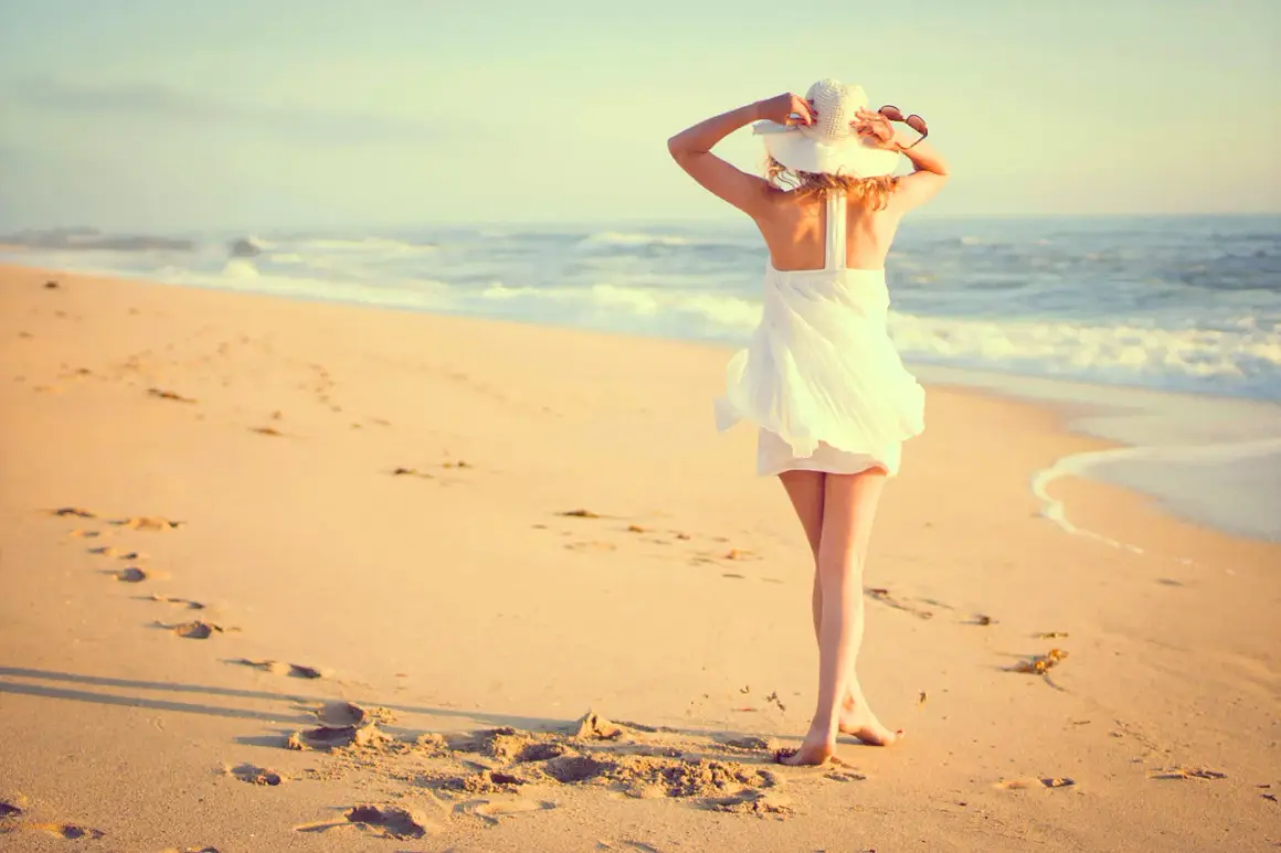 Mujer con sombrero blanco y vestido veraniego en la playa, disfrutando del mar. Estas fotos originales capturan la esencia del verano.