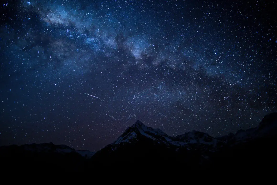 Una estrella fugaz cruza el cielo nocturno estrellado, con la Vía Láctea visible sobre montañas nevadas. Fotos perseidas capturan la magia.