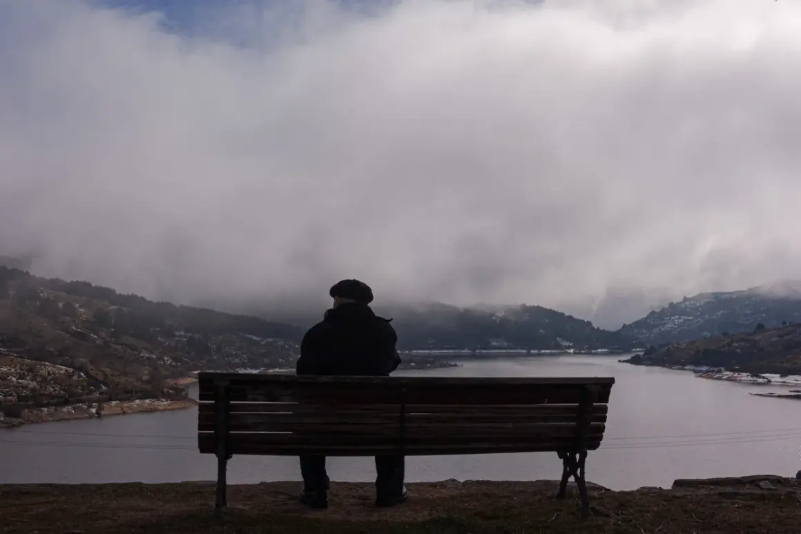 Hombre sentado en un banco, contemplando un lago y montañas cubiertas de niebla. La figura y el fondo crean una escena de introspección.