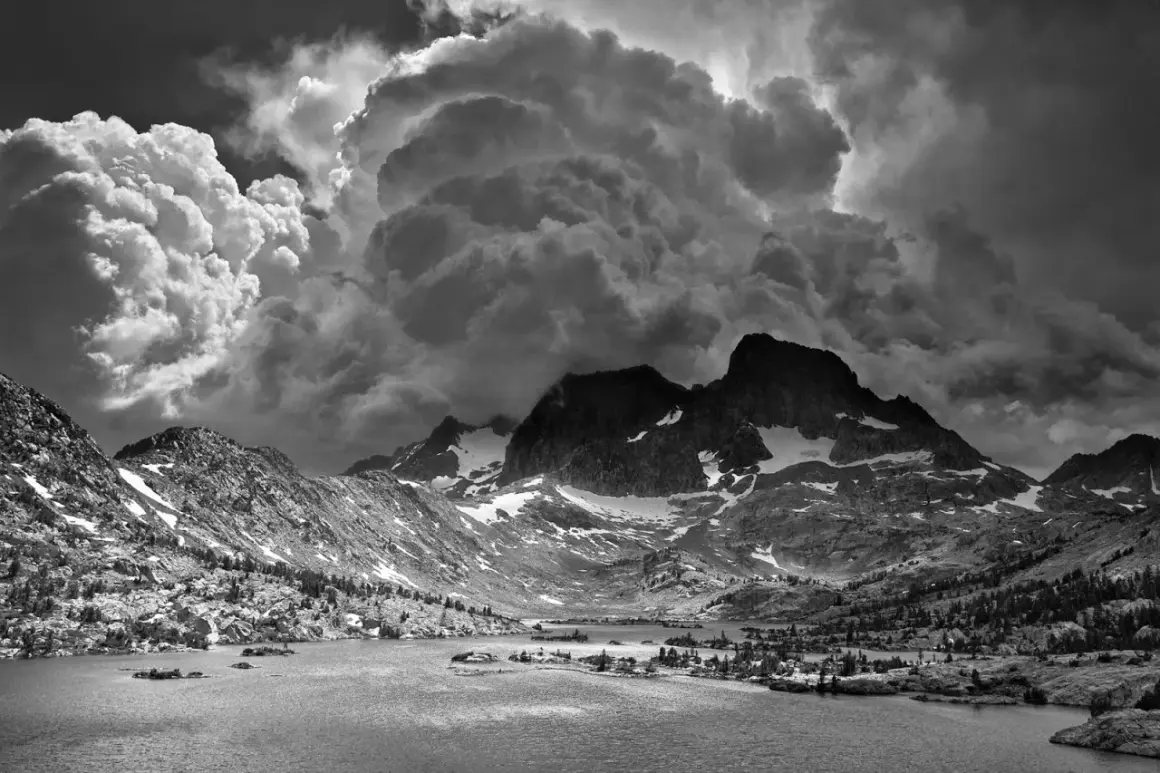 Paisaje montañoso dramático con nubes imponentes, típico de las ansel adams fotos. Un lago sereno refleja las cumbres nevadas.