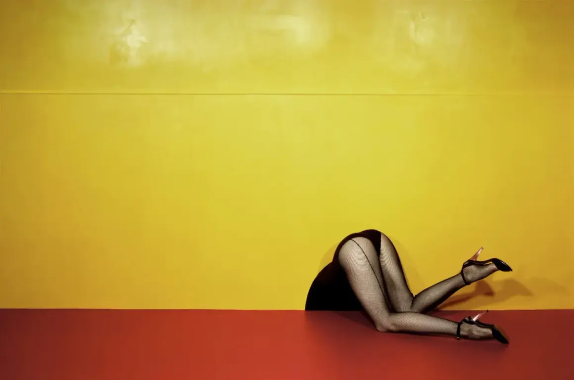 Piernas con medias de rejilla y tacones, estilo Guy Bourdin, asomando por un hueco contra un fondo amarillo y rojo.