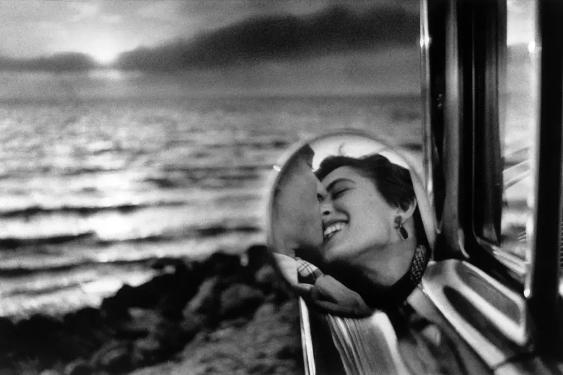 Una mujer sonríe en el espejo retrovisor de un coche, reflejando un beso. Elliott Erwitt capturó este momento íntimo junto al mar.