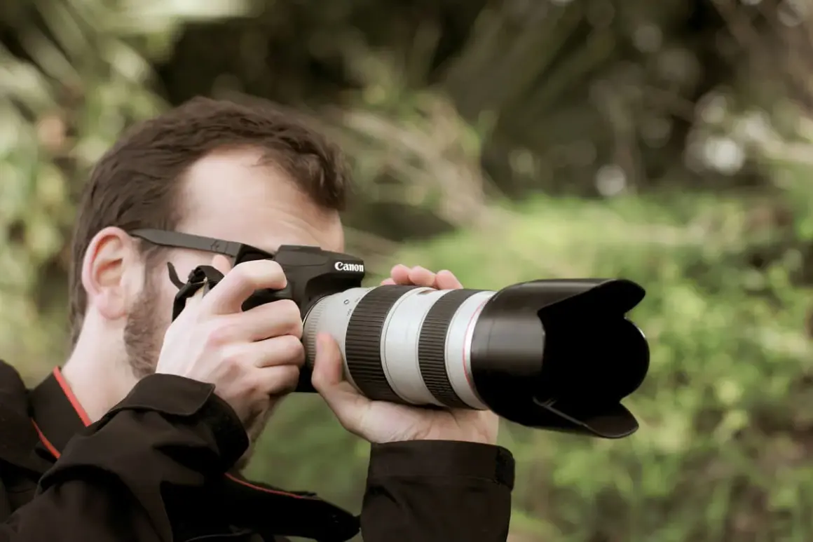 Hombre con gafas y barba toma una foto con una cámara Canon. El parasol de la lente protege de la luz.