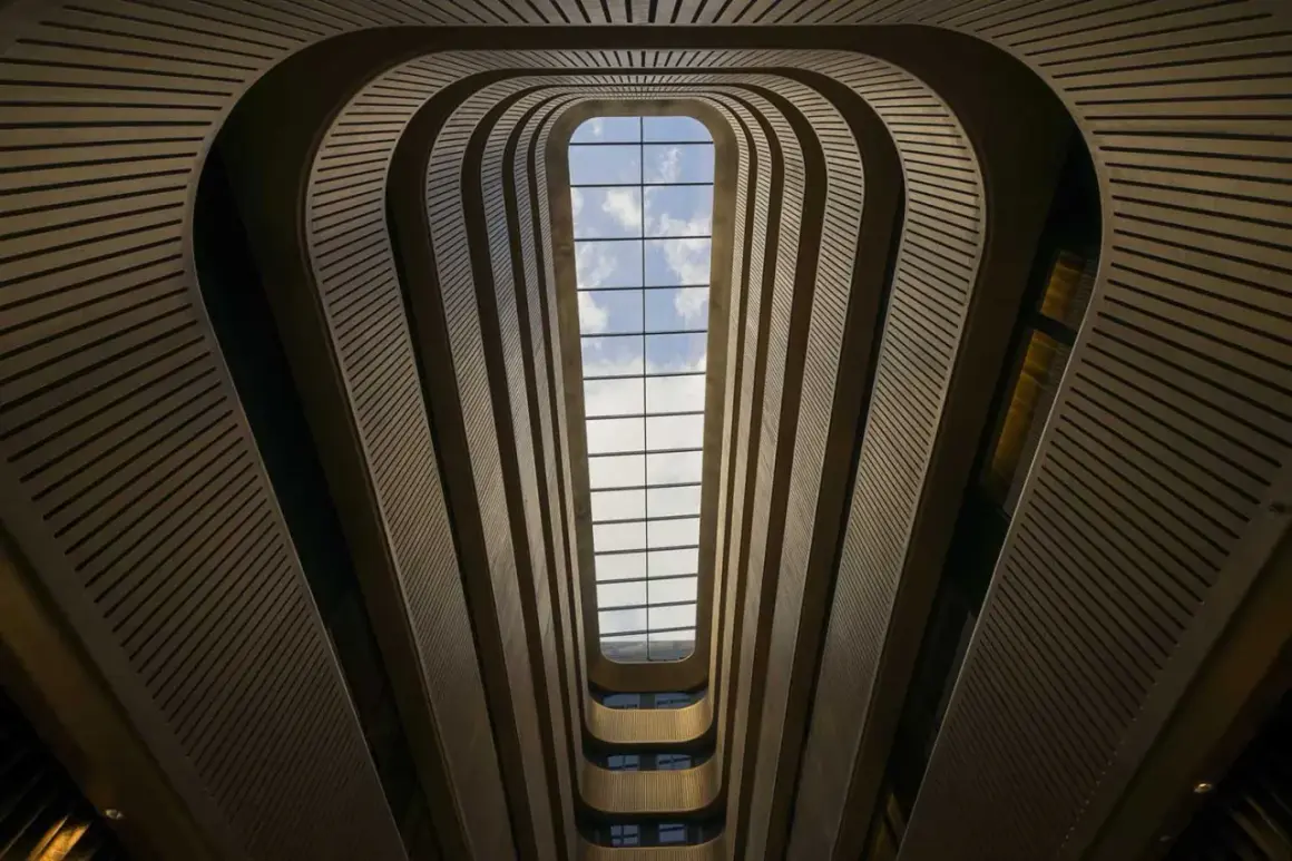 Interior de un edificio con un tragaluz rectangular. Las paredes curvas y las barandillas crean un efecto de túnel, jugando con formas geométricas básicas.