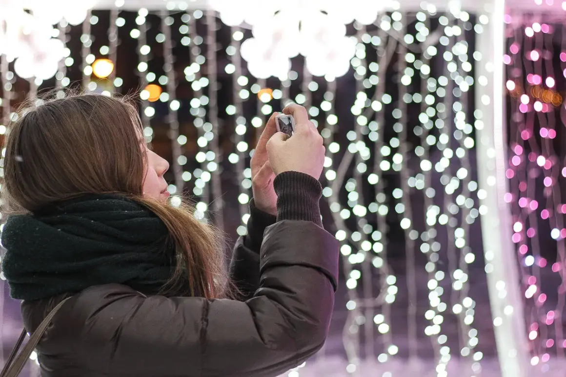 Chica con bufanda oscura toma foto de luces navideñas con un hermoso efecto bokeh.