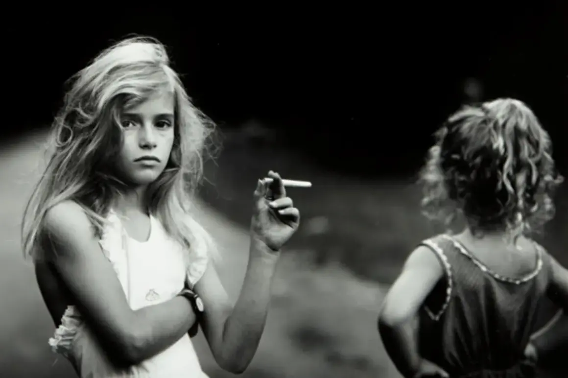Una niña rubia, en una de las sally mann fotos, sostiene un cigarrillo. Otra niña, de espaldas, mira hacia la derecha.