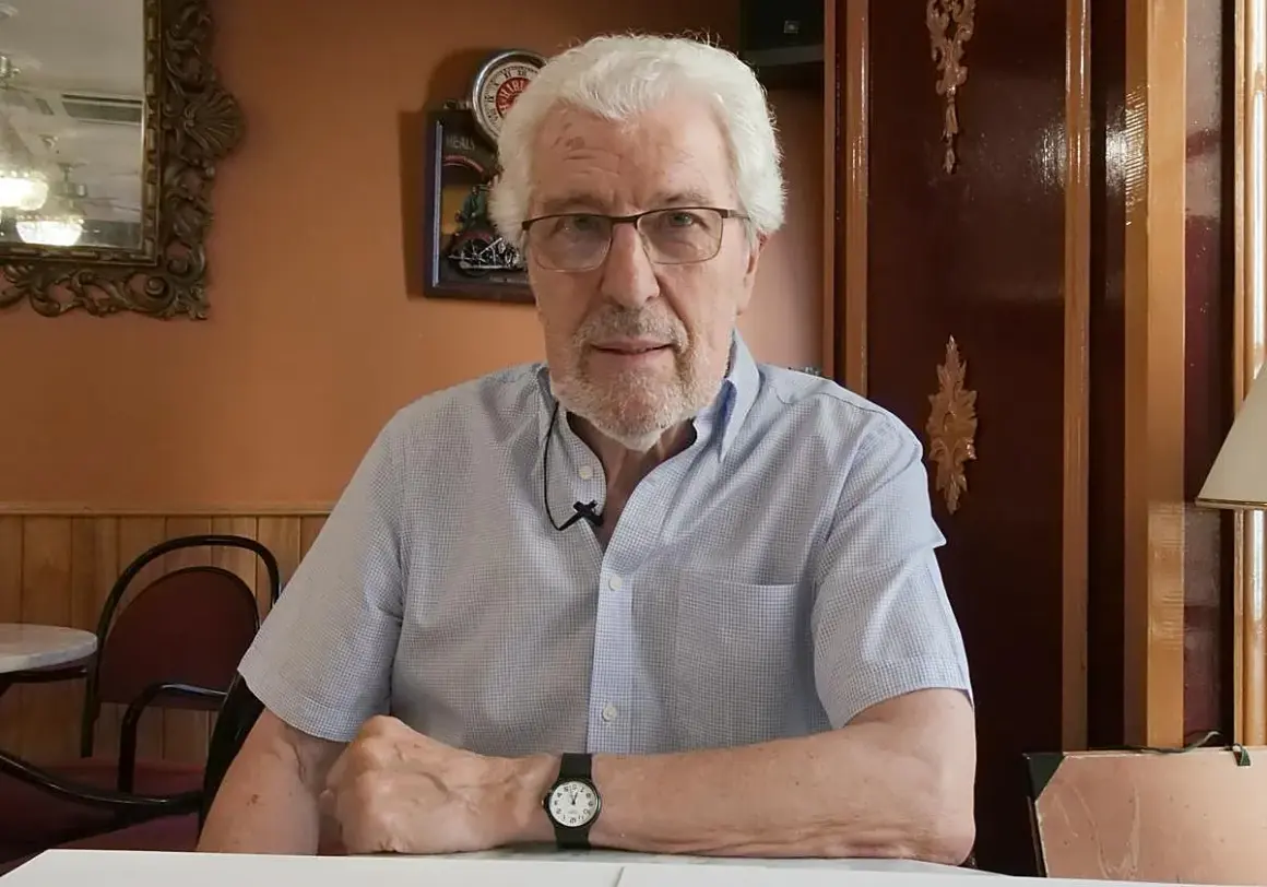 Miguel Soria, un hombre mayor con cabello blanco y gafas, sentado en un café con paredes naranjas y decoración clásica.