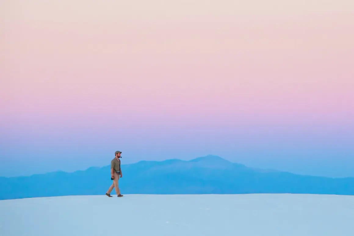 Hombre camina sobre dunas blancas bajo un cielo degradado de rosa y azul, respetando la ley del horizonte.