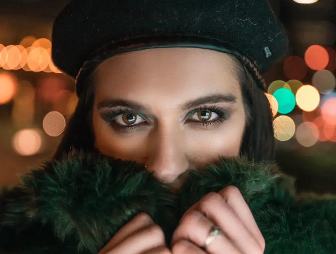Ojos penetrantes de mujer con boina negra y abrigo de pelo verde, rodeada de luces bokeh. Un reto fotográfico nocturno.