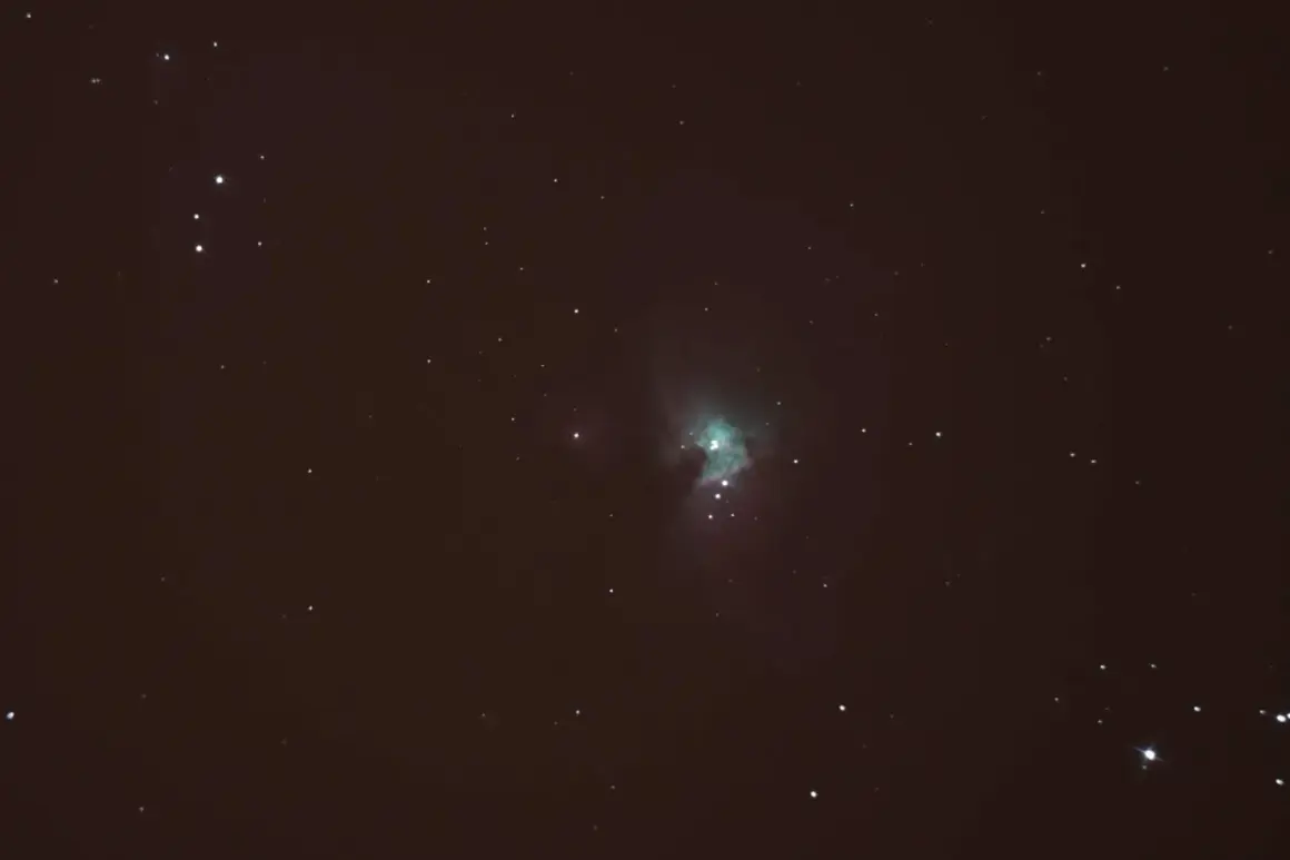 Nebulosa verdosa en el cielo nocturno, rodeada de estrellas y con tenues estelas de estrellas.