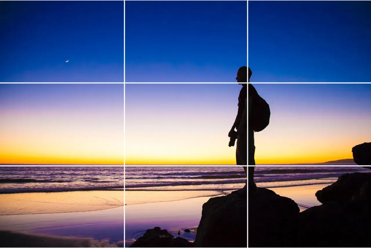 Silueta de fotógrafo en la playa al atardecer, siguiendo la regla de los tercios vertical. Luna creciente en el cielo azul.