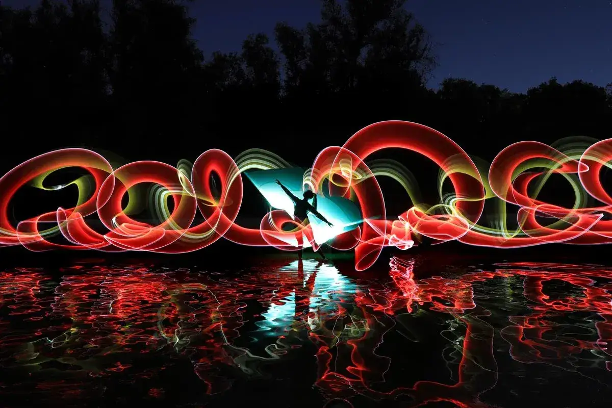 Silueta de una persona bailando con luces rojas y amarillas creando un efecto foto movida. El reflejo en el agua intensifica la magia.