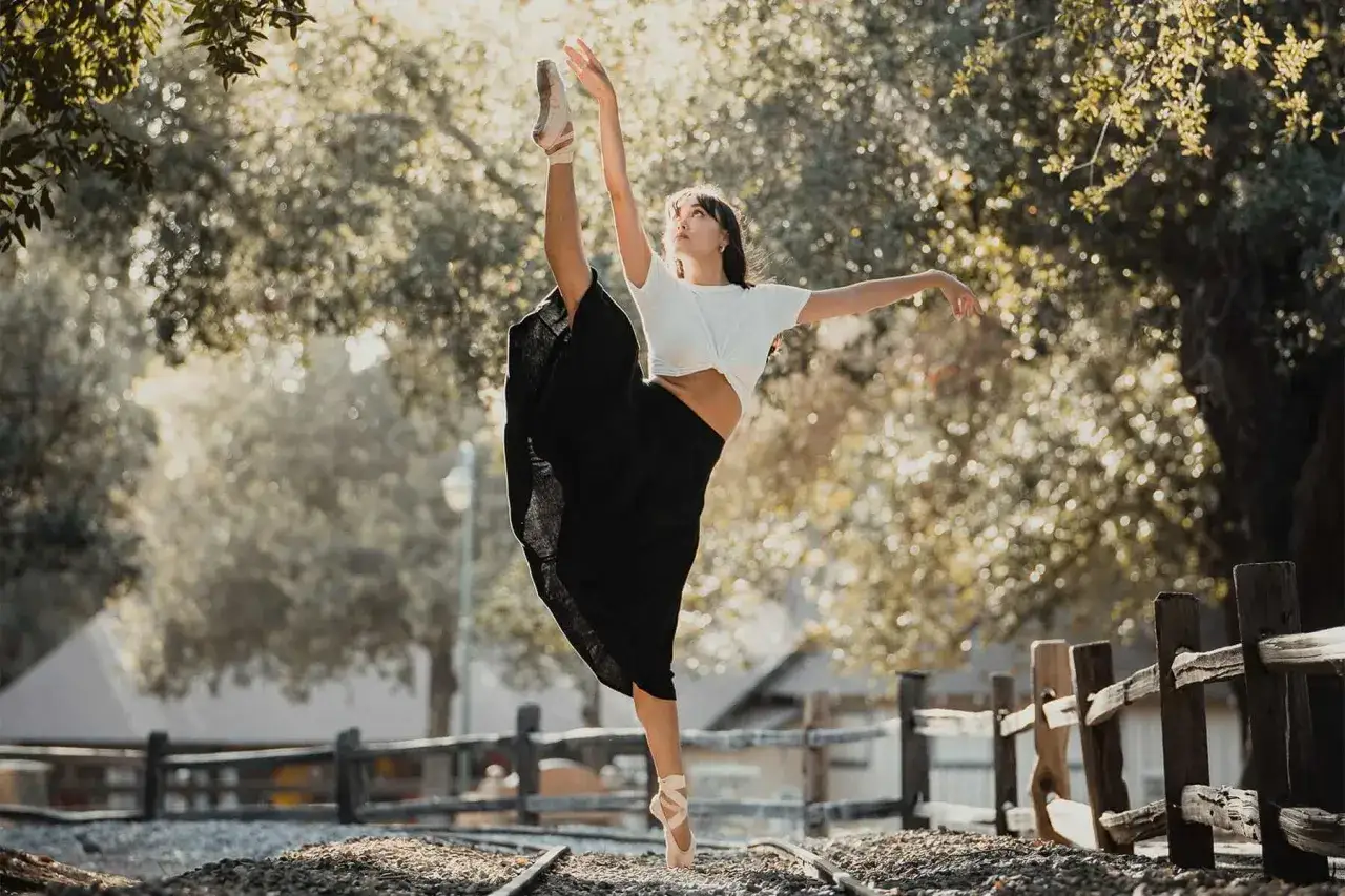 Bailarina en puntas, con pierna extendida hacia arriba, parece congelar fotograf&iacute;a en un entorno natural con v&iacute;as de tren.
