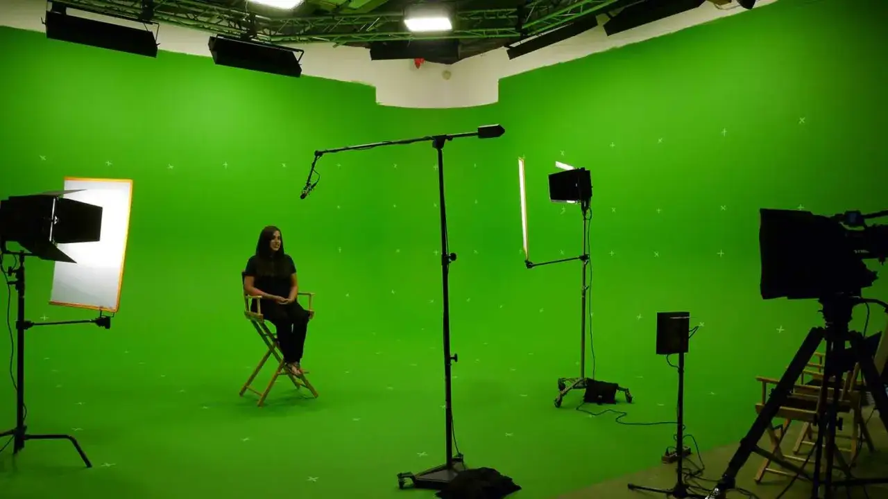 Estudio de croma que es un fondo verde para efectos visuales. Una mujer sentada en una silla de director, rodeada de equipo de filmaci&oacute;n.