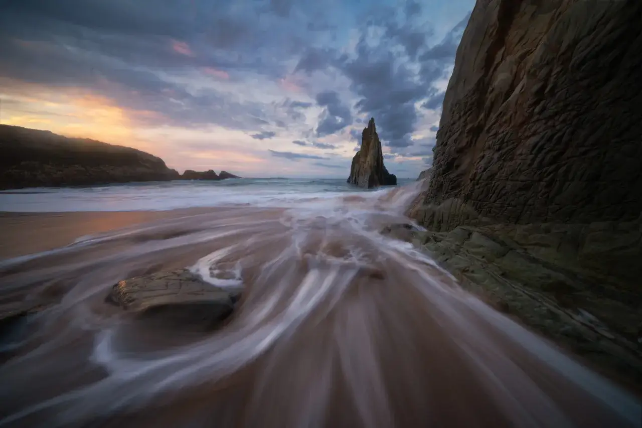 Olas sedosas fluyen sobre la arena en esta fotograf&iacute;a de larga exposici&oacute;n, con un pin&aacute;culo rocoso solitario emergiendo del mar al atardecer.