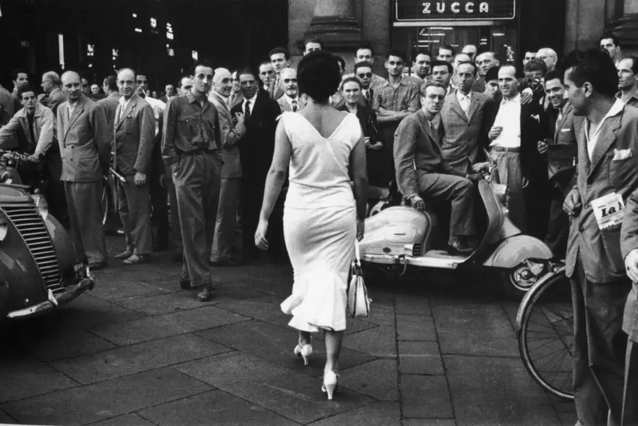 Evgen Bavcar captura una mujer de espaldas, vestida de blanco, caminando entre una multitud de hombres. Una Vespa y un coche clásico completan la escena.