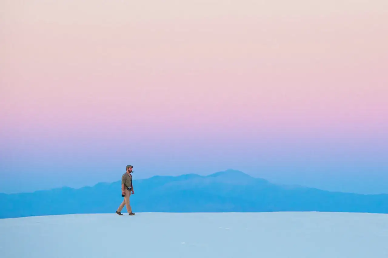 Hombre camina sobre dunas blancas bajo un cielo degradado de rosa y azul, respetando la ley del horizonte.