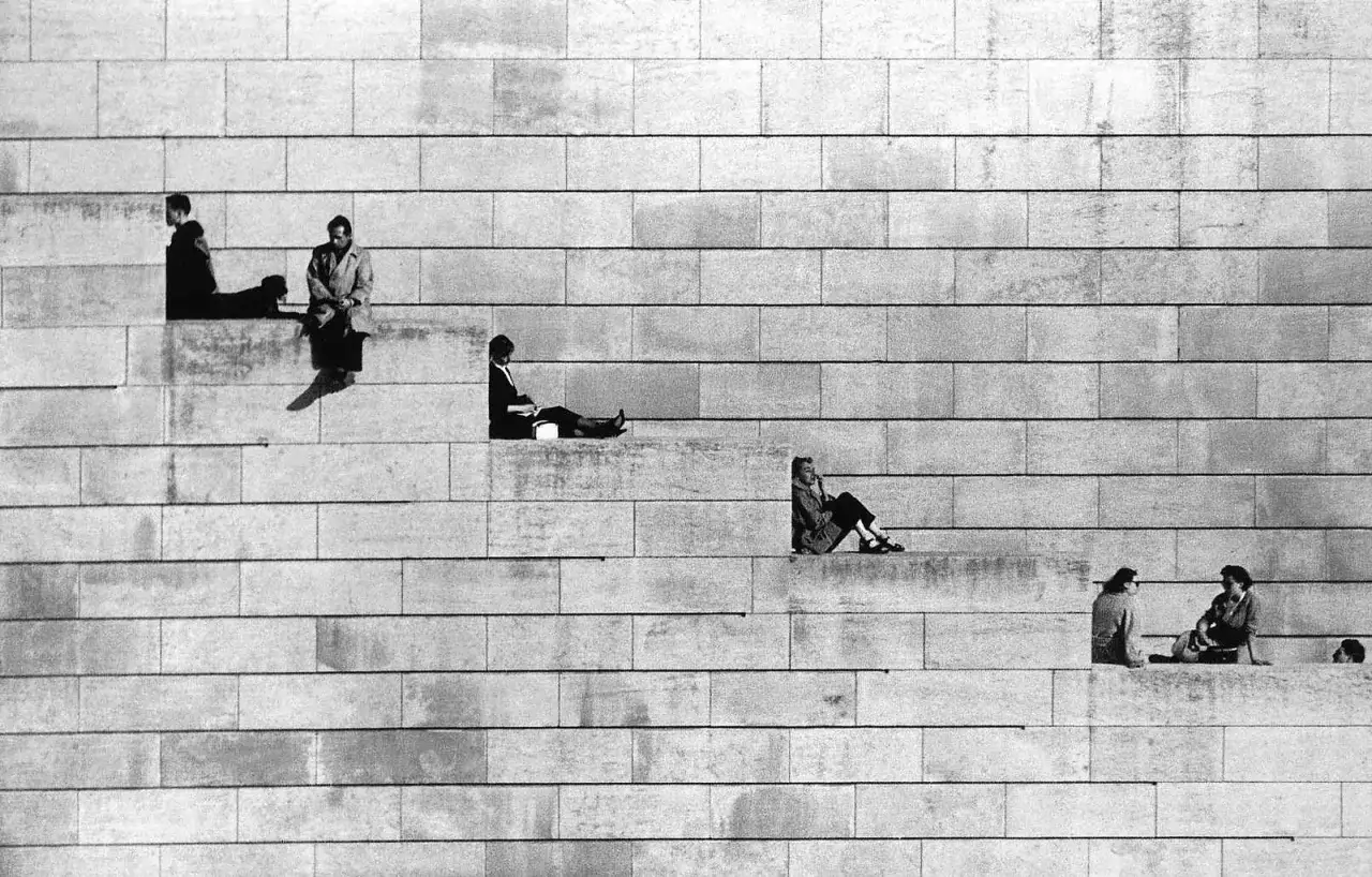 Personas sentadas en escalones de piedra, creando una línea horizontal de descanso y contemplación contra una pared texturizada.