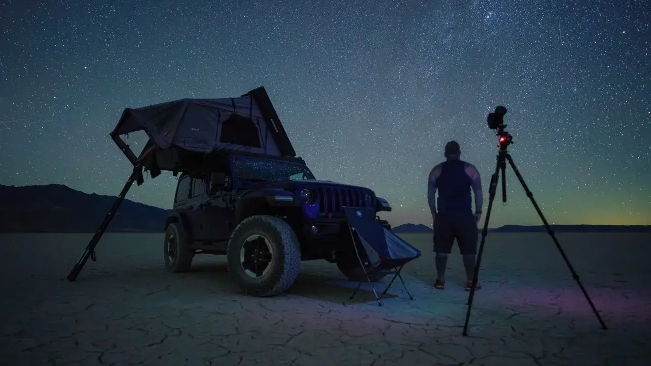 Un hombre observa un cielo estrellado en una fotografía nocturna. Un Jeep con tienda de techo y una cámara en trípode completan la escena.