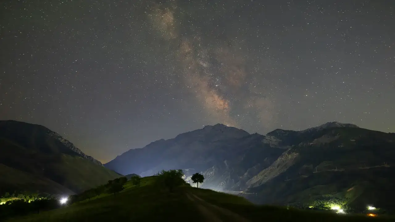 Un camino de tierra atraviesa una colina verde hacia un &aacute;rbol solitario bajo la v&iacute;a l&aacute;ctea en el cielo. Monta&ntilde;as imponentes se alzan en la oscuridad.