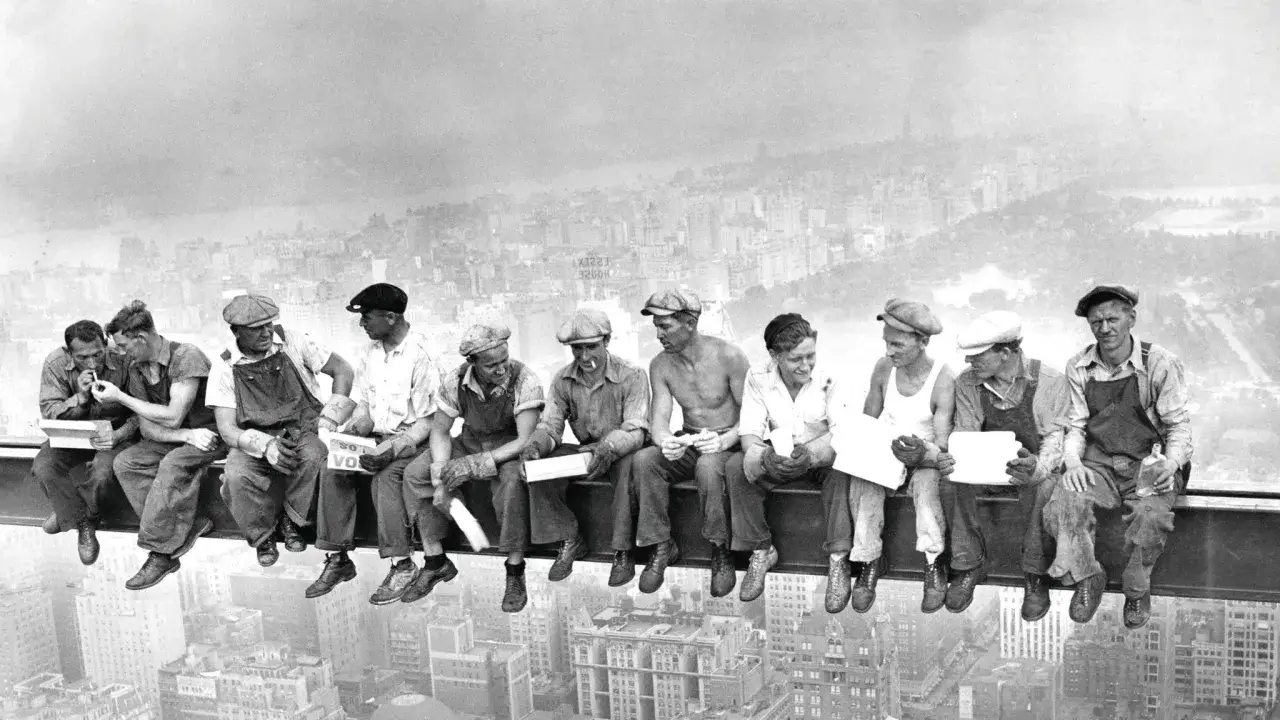 foto obreros nueva york: once trabajadores descansan en una viga de acero, con la ciudad de Nueva York de fondo.