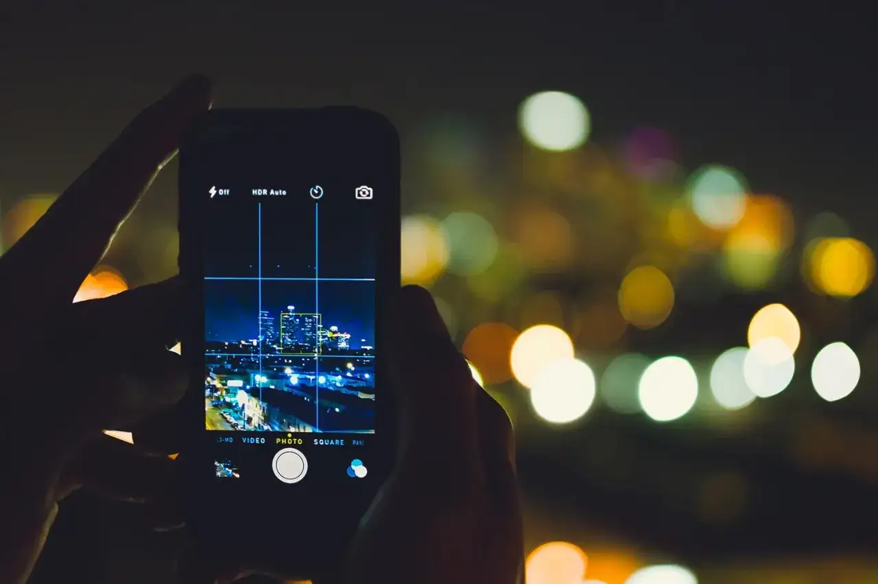Persona capturando la silueta de una ciudad nocturna, luces bokeh de fondo, mientras intenta hacer fotos con el móvil.