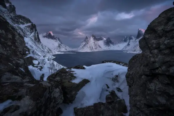 Foto panoramica de montañas nevadas y un lago oscuro bajo un cielo tormentoso. La luz del amanecer tiñe de rosa las cumbres.