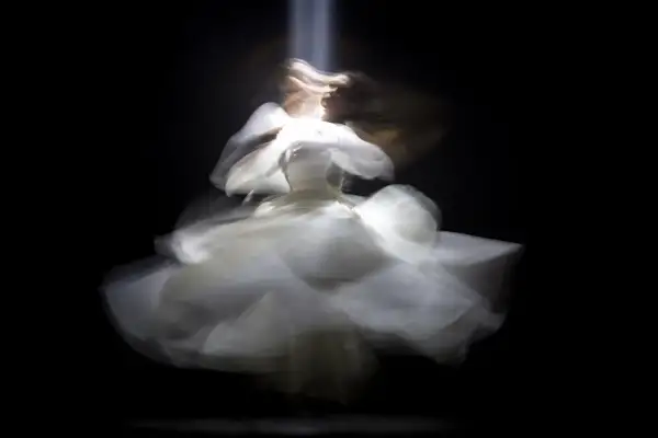 Figura etérea con un vestido blanco vaporoso, capturada con un **efecto foto movida** que evoca un baile onírico bajo un haz de luz.