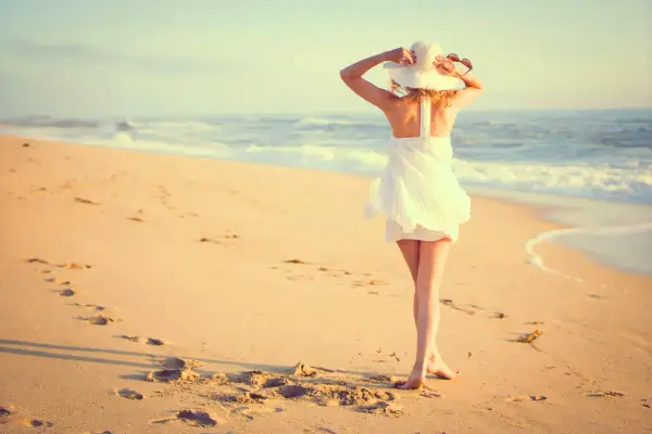 Mujer con sombrero blanco y vestido veraniego en la playa, disfrutando del mar. Estas fotos originales capturan la esencia del verano.