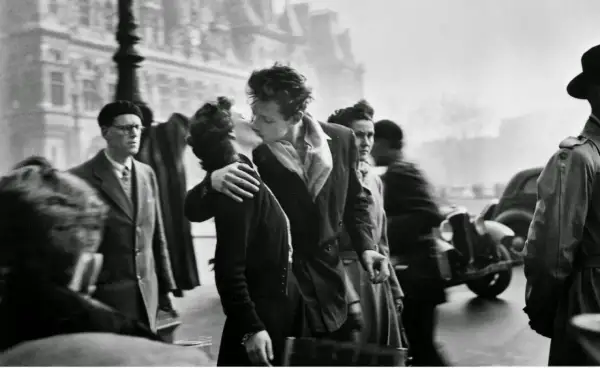Pareja besándose apasionadamente en París, rodeada de gente y un coche antiguo. Un momento capturado en estas fotos históricas.