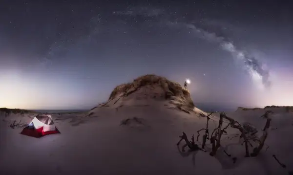 Persona ilumina la noche estrellada con su linterna, cerca de una tienda de campaña en la playa. Las fotos con ruido capturan la magia del cielo nocturno.