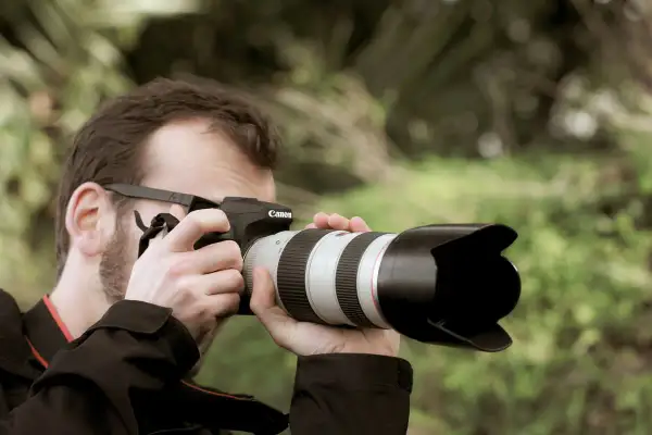 Hombre con gafas y barba toma una foto con una cámara Canon. El parasol de la lente protege de la luz.