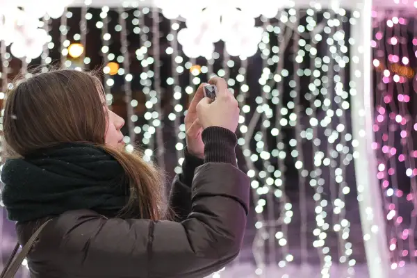 Chica con bufanda oscura toma foto de luces navideñas con un hermoso efecto bokeh.