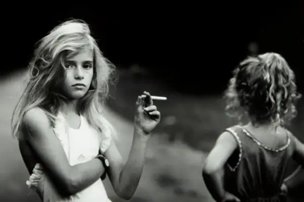 Una niña rubia, en una de las sally mann fotos, sostiene un cigarrillo. Otra niña, de espaldas, mira hacia la derecha.