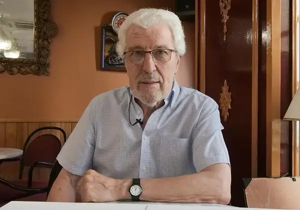 Miguel Soria, un hombre mayor con cabello blanco y gafas, sentado en un café con paredes naranjas y decoración clásica.