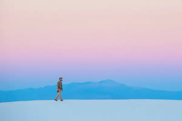 Hombre camina sobre dunas blancas bajo un cielo degradado de rosa y azul, respetando la ley del horizonte.